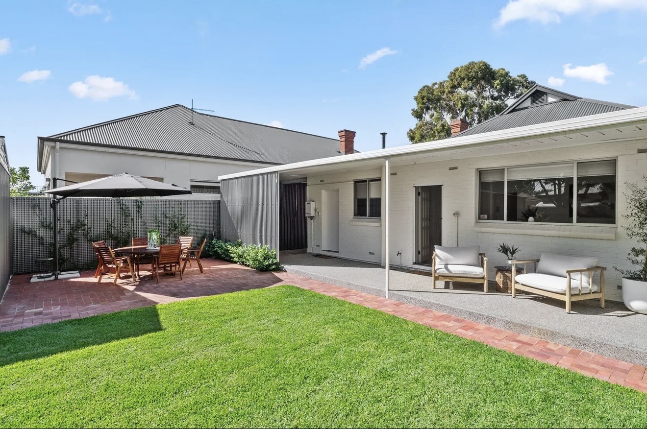 Yard Theory backyard with a green lawn, a brick-paved dining patio under a charcoal umbrella, and a covered walkway with lounge chairs beside a charcoal screen